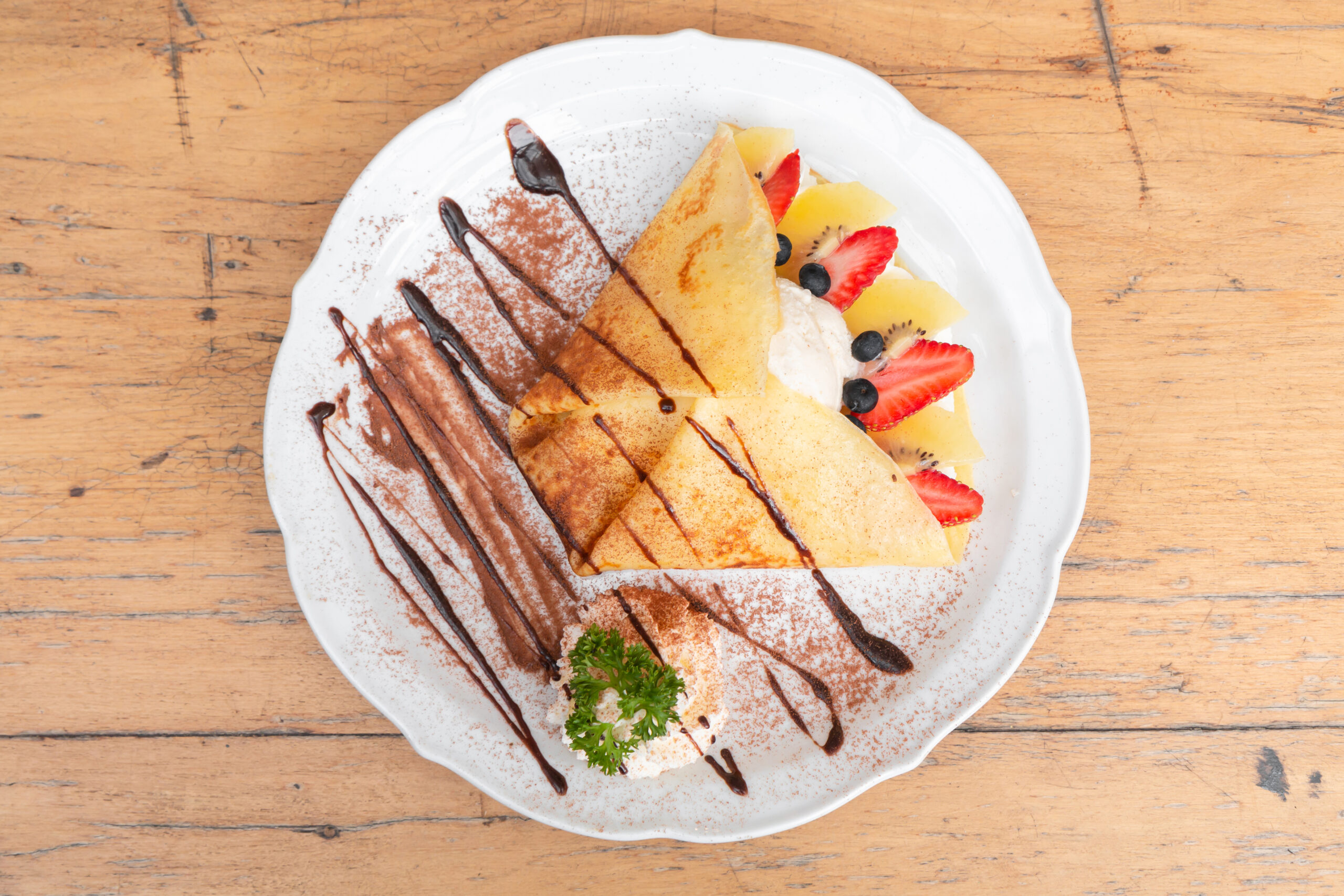 top view of cold crepe with vanilla ice cream, strawberry, kiwi and whipped cream topped with chocolate sauce on the wooden table.